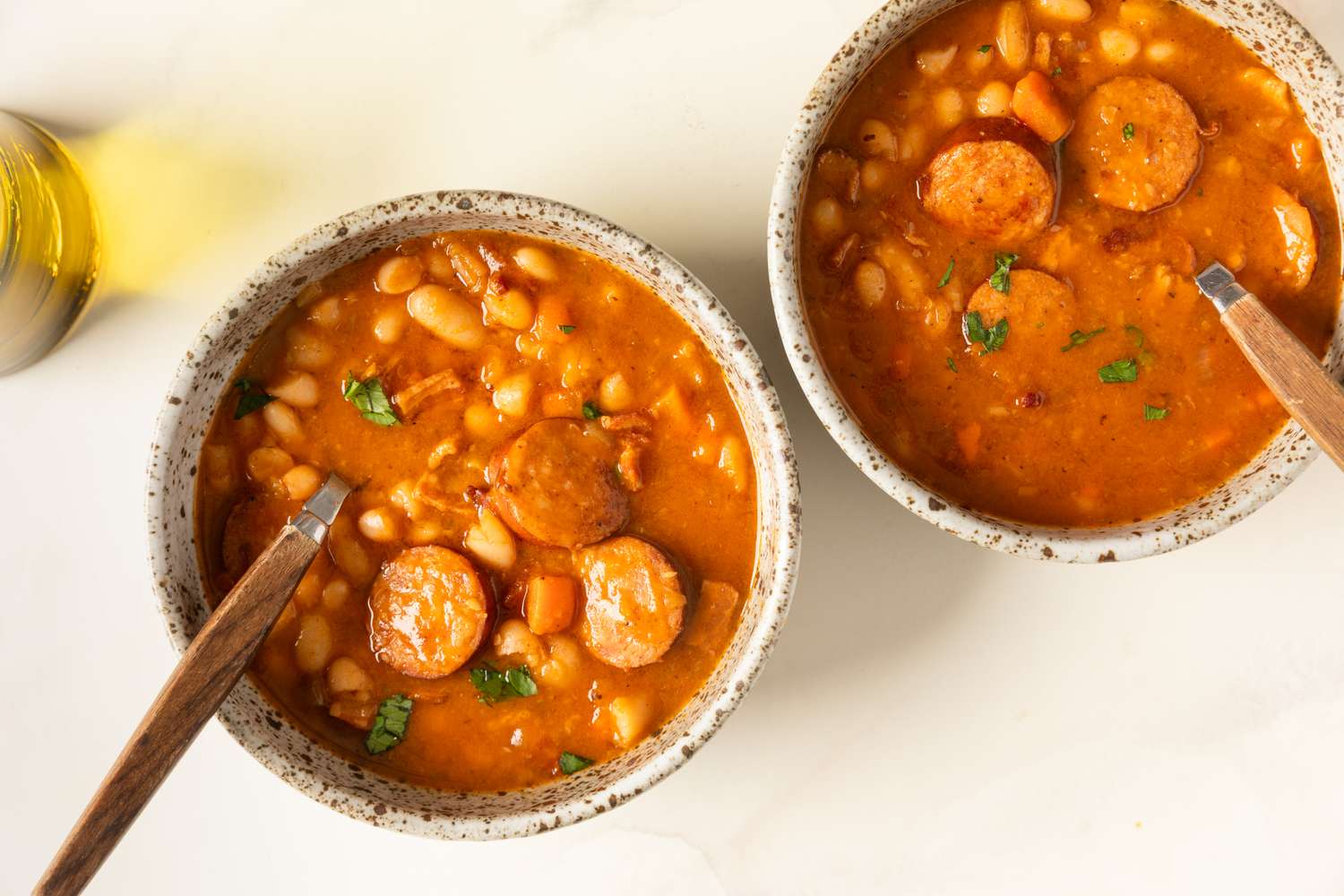 Two bowls of bean and sausage stew on a white surface wooden spoons in each bowl