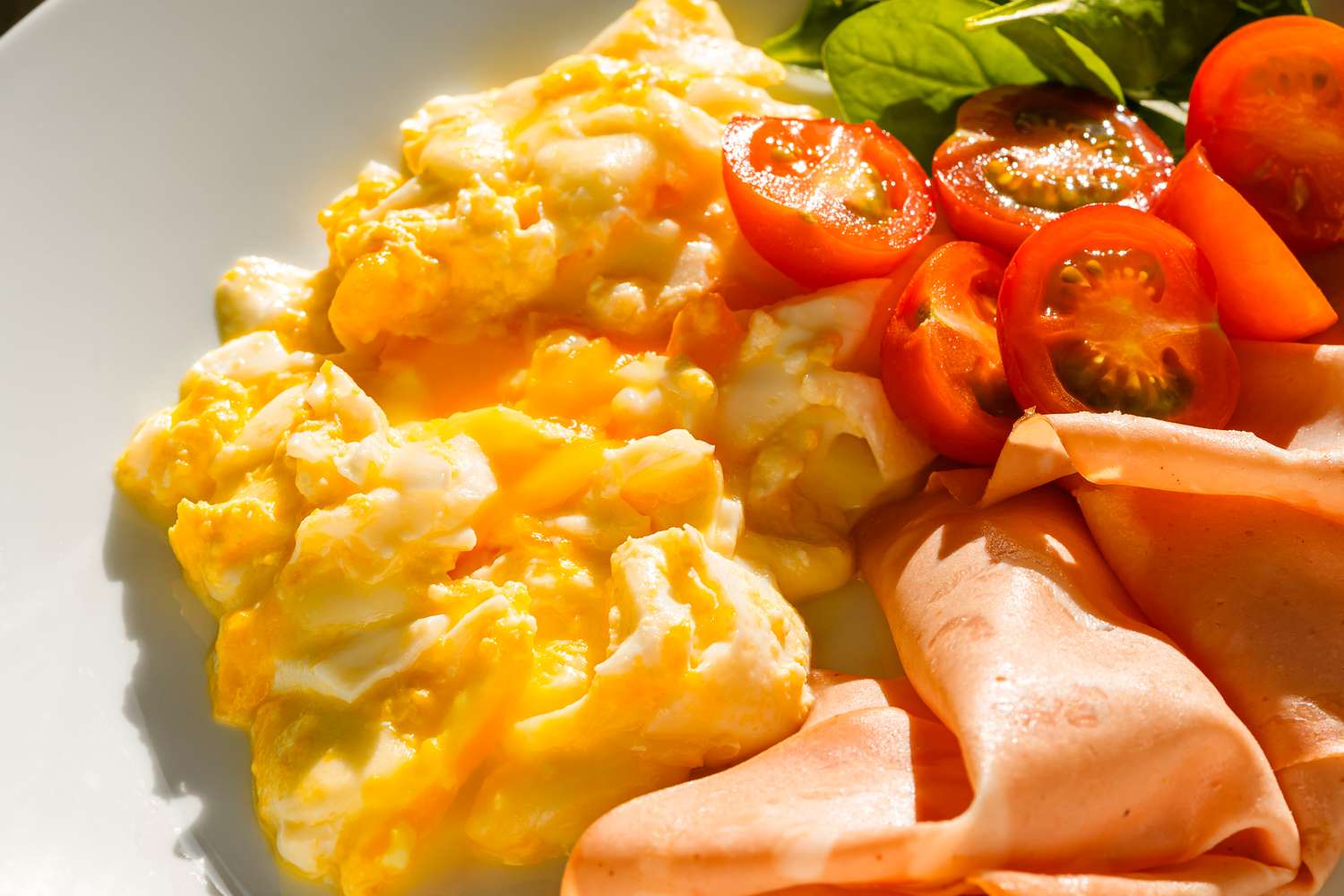 Scrambled eggs served with sliced tomatoes greens and deli meat on a plate