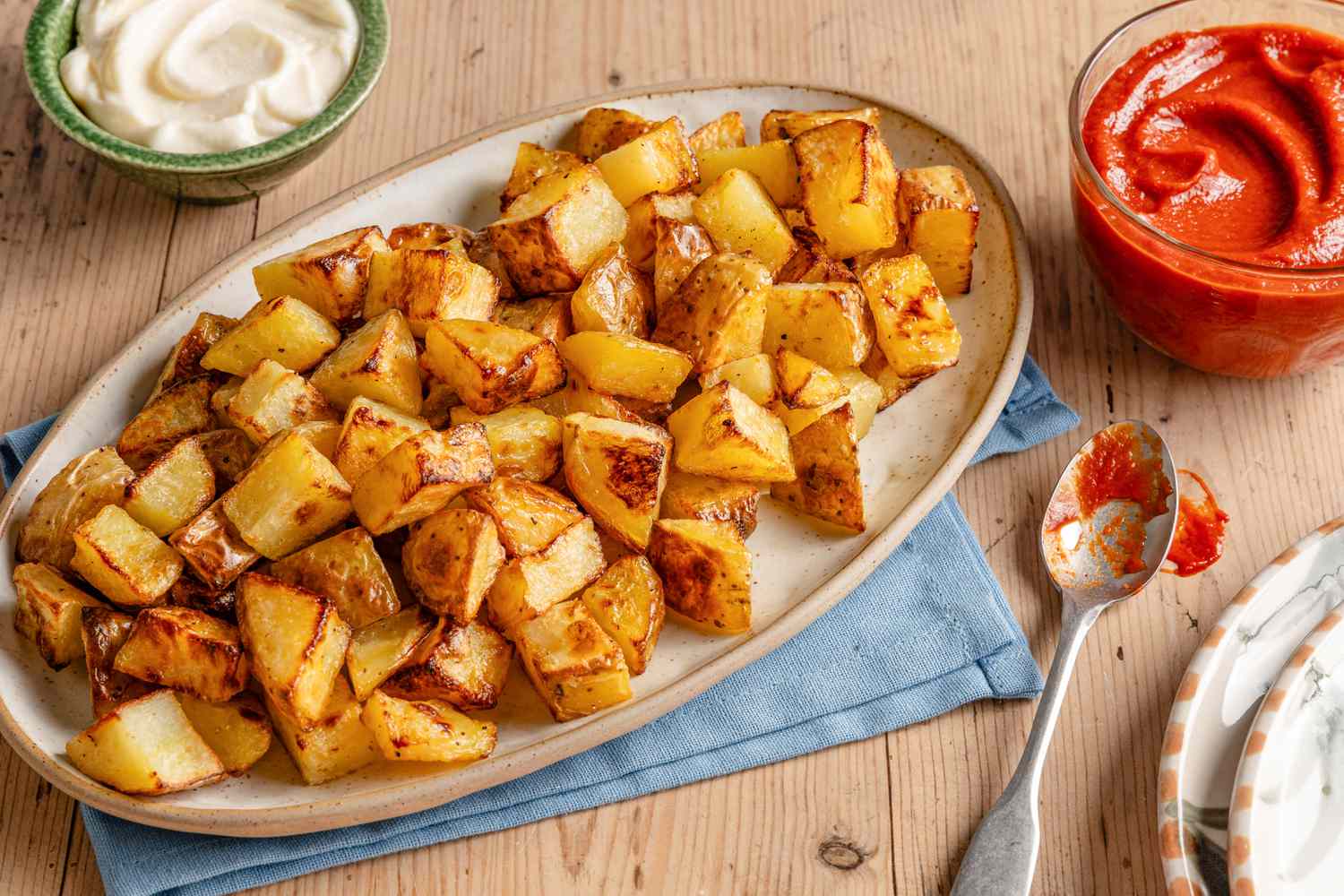 Angled view of a platter of patatas bravas next to a spoon, and bowls of tomato sauce and aioli all on a wooden tabletop