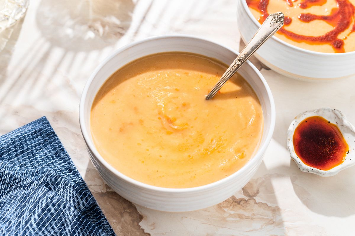Angled view of a white bowl of cauliflower soup next to a small bowl of paprika oil, a blue striped napkin and two clear drinking glasses