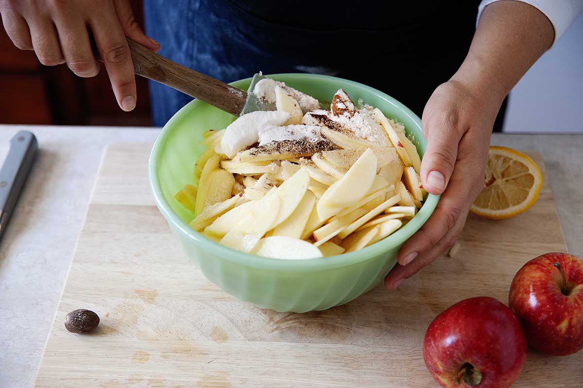Woman using a spatula to mix spices, sugar and flour into apple slices in a green bowl.