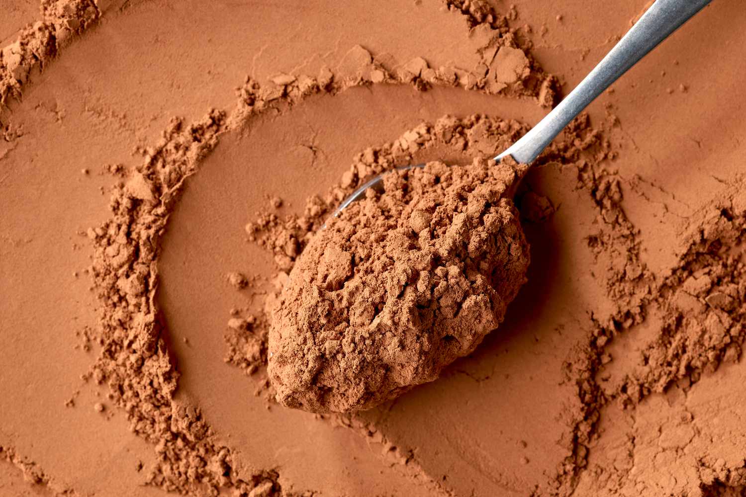 A spoon holding a scoop of powdered cocoa laid over a background of cocoa powder