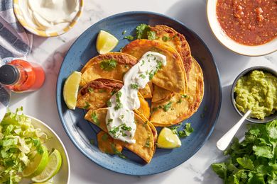 Bowl of cheesy baked tacos on a plate topped with sour cream and cilantro at a table setting with a bowl of lime wedges and shredded lettuce, bowl of sour cream, bowl of salsa, bowl of salsa, and a bunch of cilantro