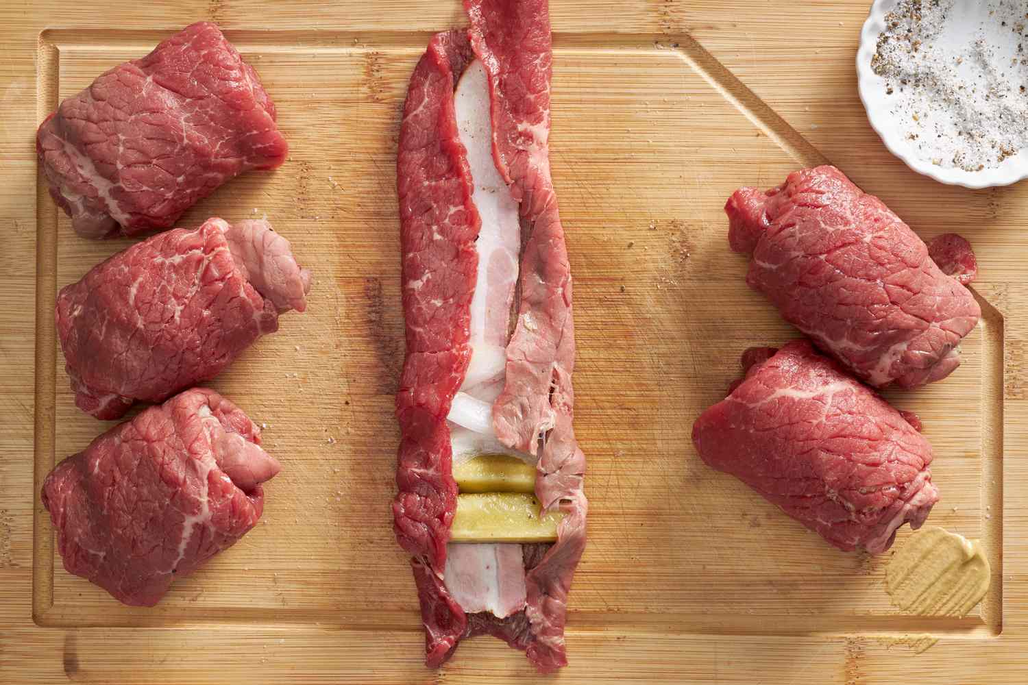 Making rouladen: edges of beef folded over the filling (on a wooden cutting board next to already rolled rouladen) 