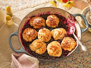 blueberry grunt in a cast iron braiser at cozy looking table with a stack of bowls, salt shakers, a napkin holder with single use napkins, and a vase of flowers