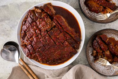 Overhead view of a white platter of sliced beef brisket next to serving spoons and two plates of brisket with forks all on a marbled countertop