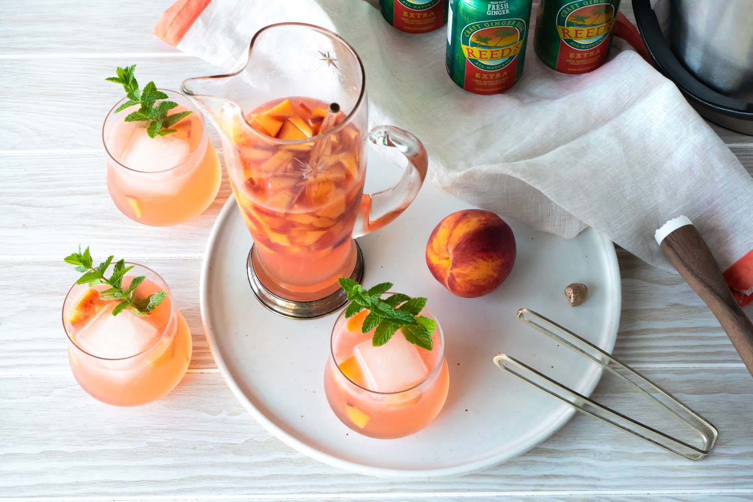 Ginger Peach Rum Punch in Three Glasses Next to a Pitcher of More Punch, Ginger Beer, Ice Tongs, and Nutmeg