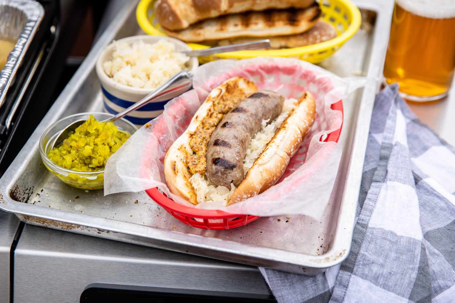 Beer Brats Topped with Onion and Mustard Next to Bowls with Onions and Relish