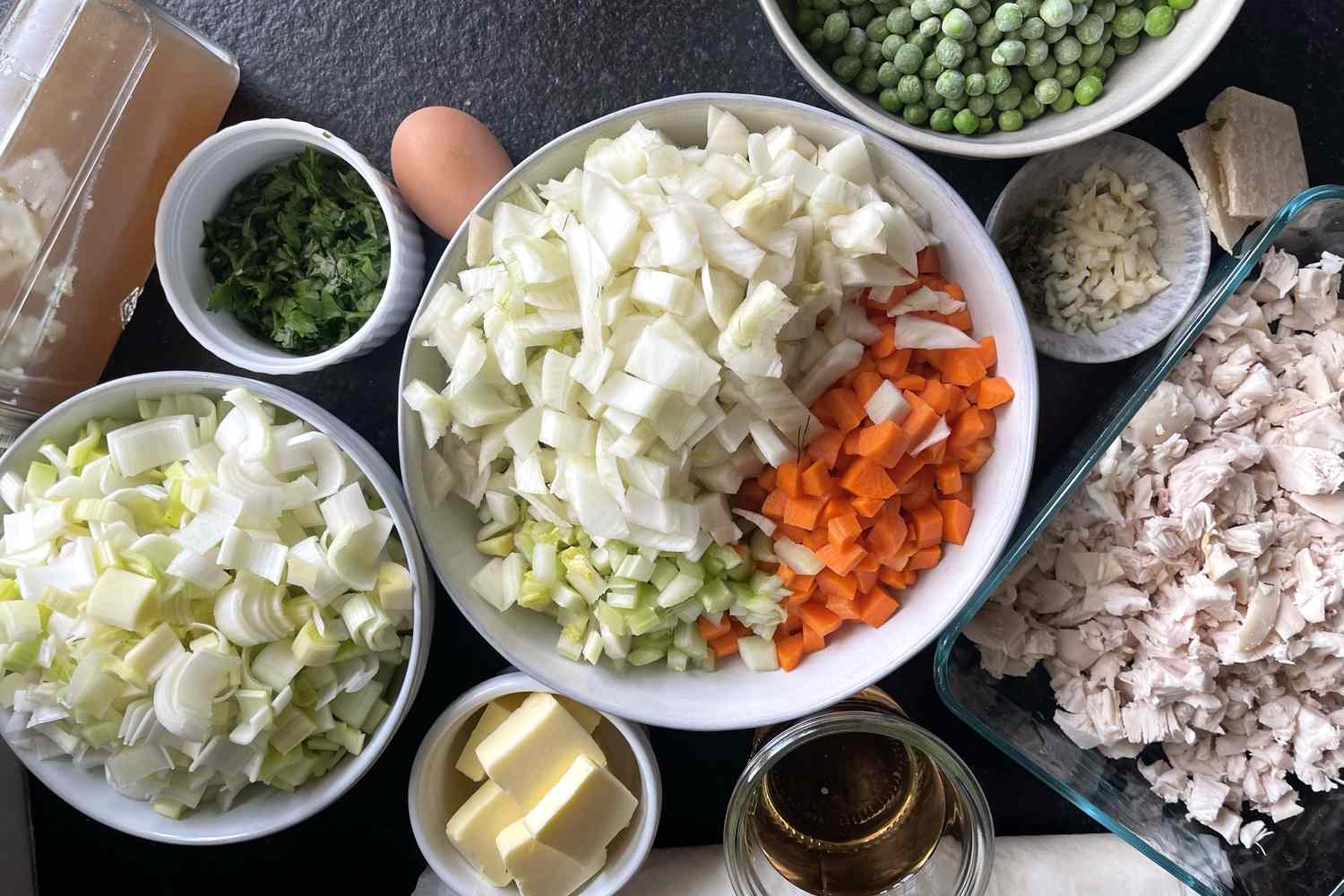 Ingredients for a chicken pot pie including diced vegetables cooked chicken and peas arranged in bowls on a dark surface