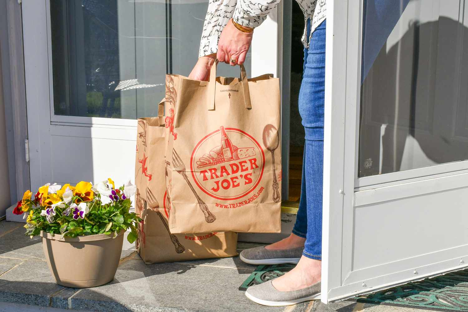 A woman wearing jeans picking up two Trader Joe's bags from a front porch to bring them inside.
