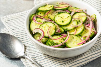 A bowl of cucumber salad with slices of cucumber and red onion on a striped cloth next to a spoon