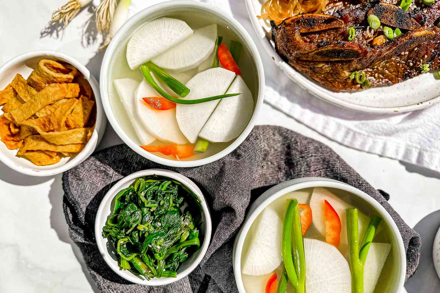 Bowl of Dongchimi Surrounded by Bowls of Banchan (Side Dishes) and a Bowl of Korean Short Ribs. On the Table, a Table Napkin and Some Whole Green Onions.