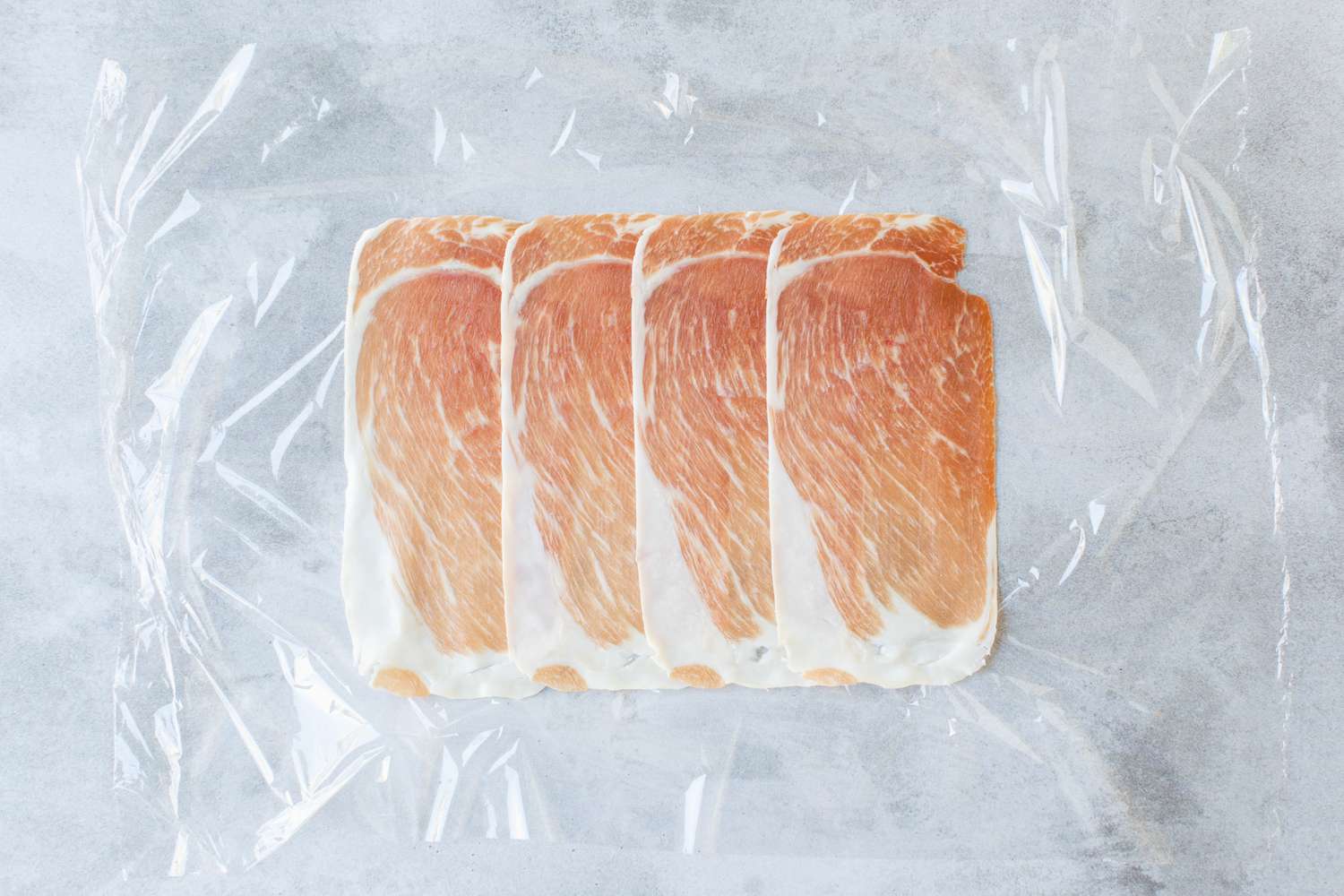 Four Slices of Prosciutto Lined up on a Piece of Plastic Wrap