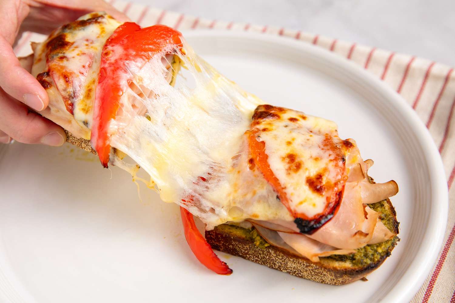 Open-Face Turkey Pesto Sandwich, cut in half with a delicious looking cheese-pull as a hand pulls half of the sandwich off of the plate