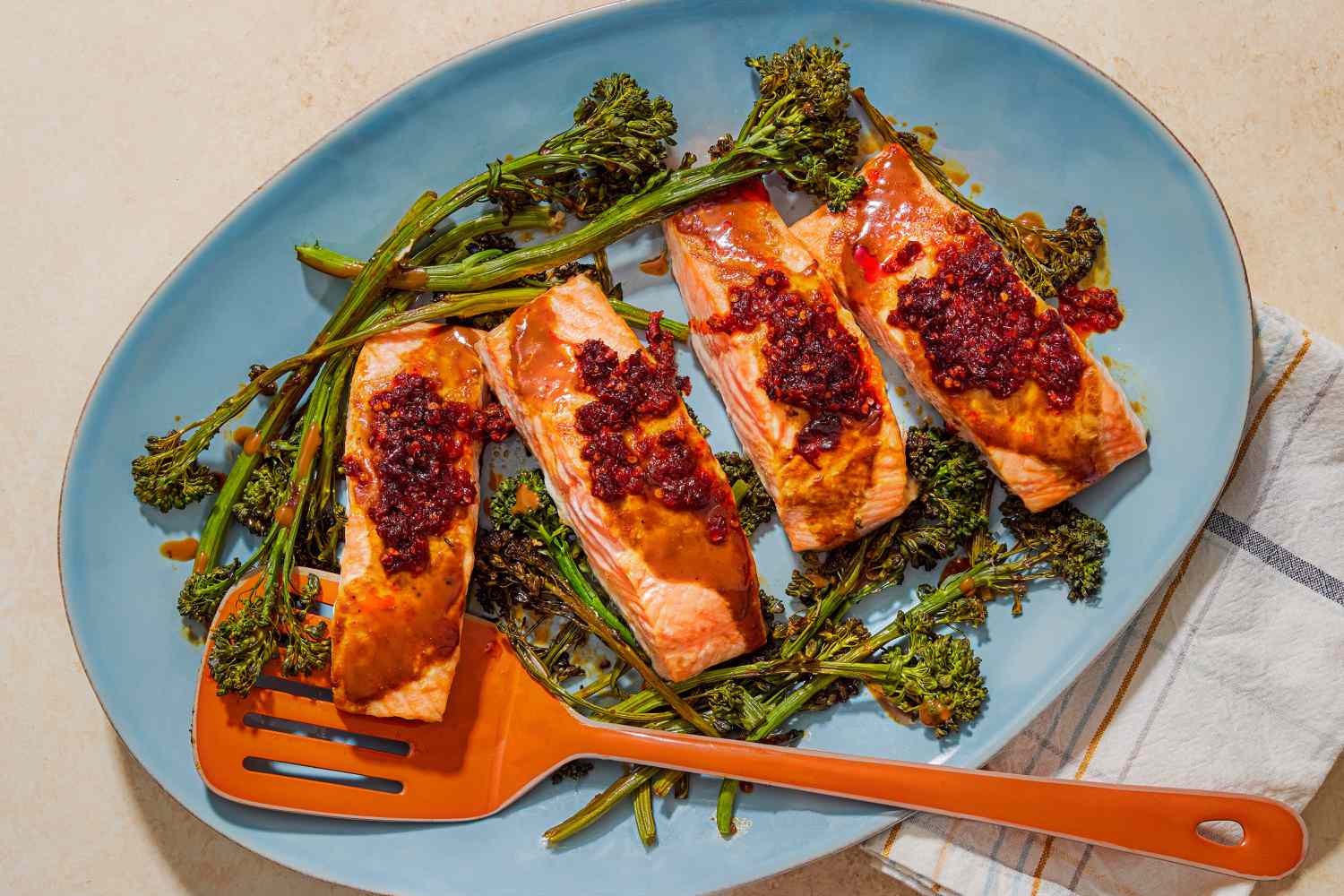 Overhead view of a blue serving platter of cooked four salmon filets topped with chili crisp and stems of broccolini with an orange spatula under a piece of salmon