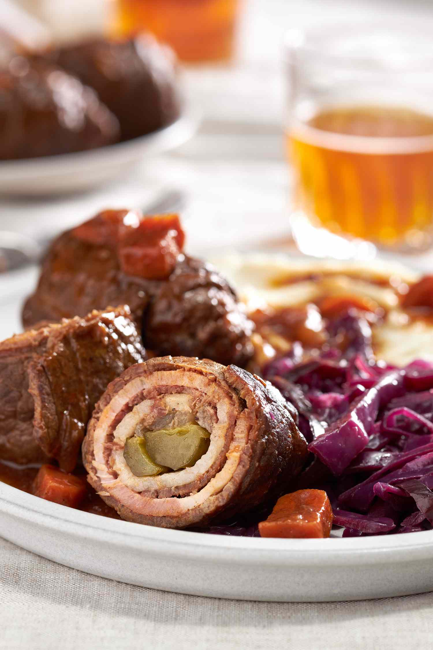German rouladen with cabbage and mashed potatoes and gravy on a plate and in the background, a table setting with another serving on a plate and a few glasses of beer