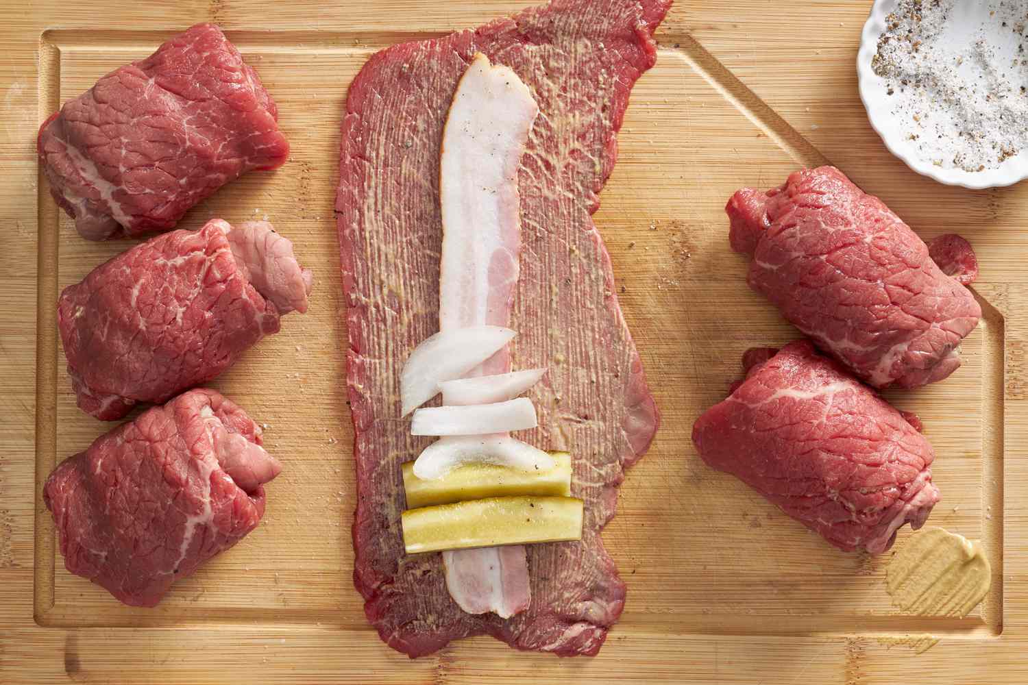 Making rouladen: bacon, pickles, and onion on a thin piece of beef (on a wooden cutting board next to already rolled rouladen) 