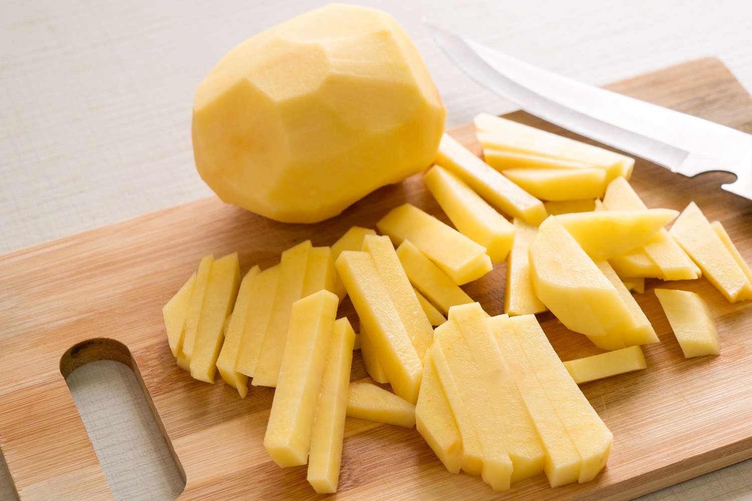 A peeled potato and sliced up potatoes on a wooden cutting board, with a knife on the right side