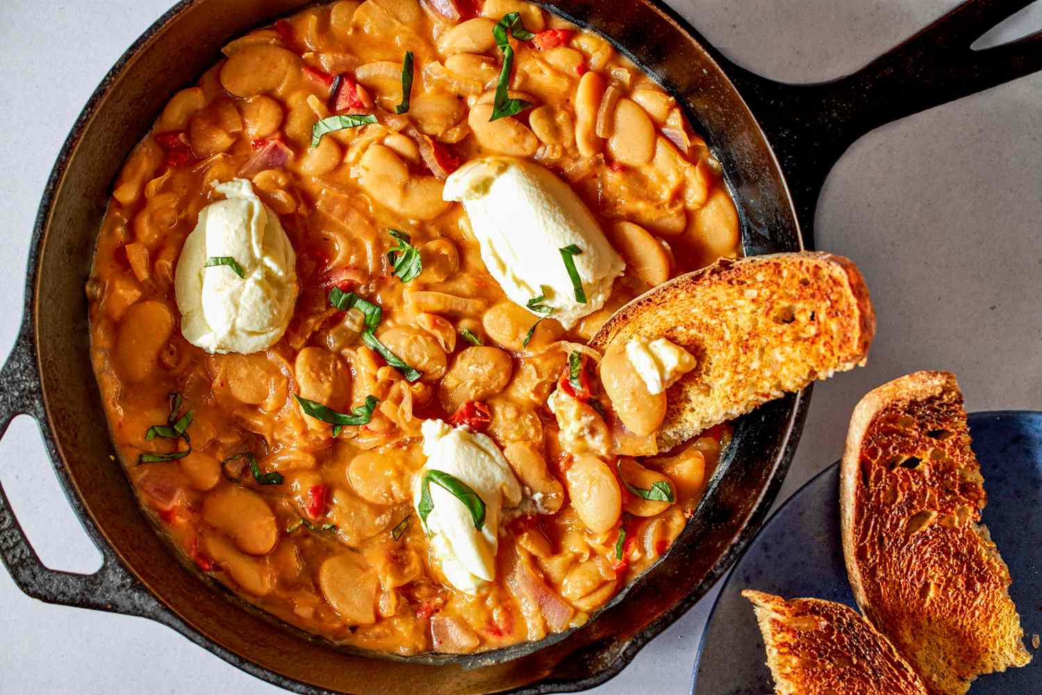 Overhead shot of Tuscan butter beans in a skillet topped with dollops of ricotta toasted bread on the side