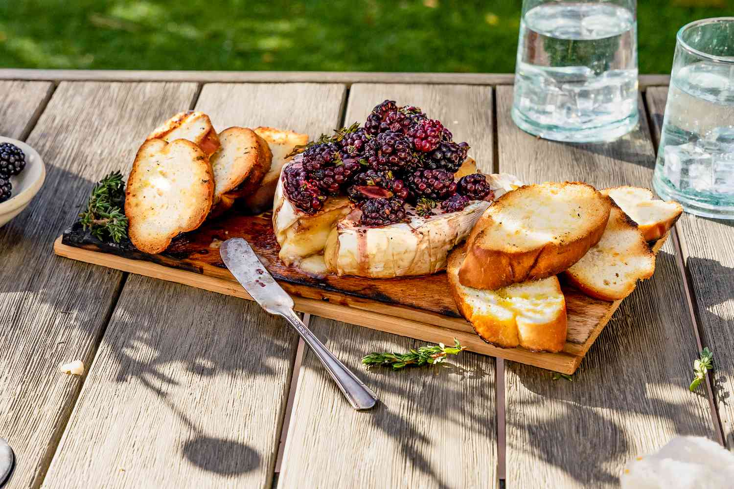 Picnic Table Setting: Cedar Plank Grilled Brie Topped With Black Berries and a Sprig of Thyme, on the Plank Next to the Brie, Slices of Baguette, and on the Table, Two Glasses of Water and Sprigs of More Thyme