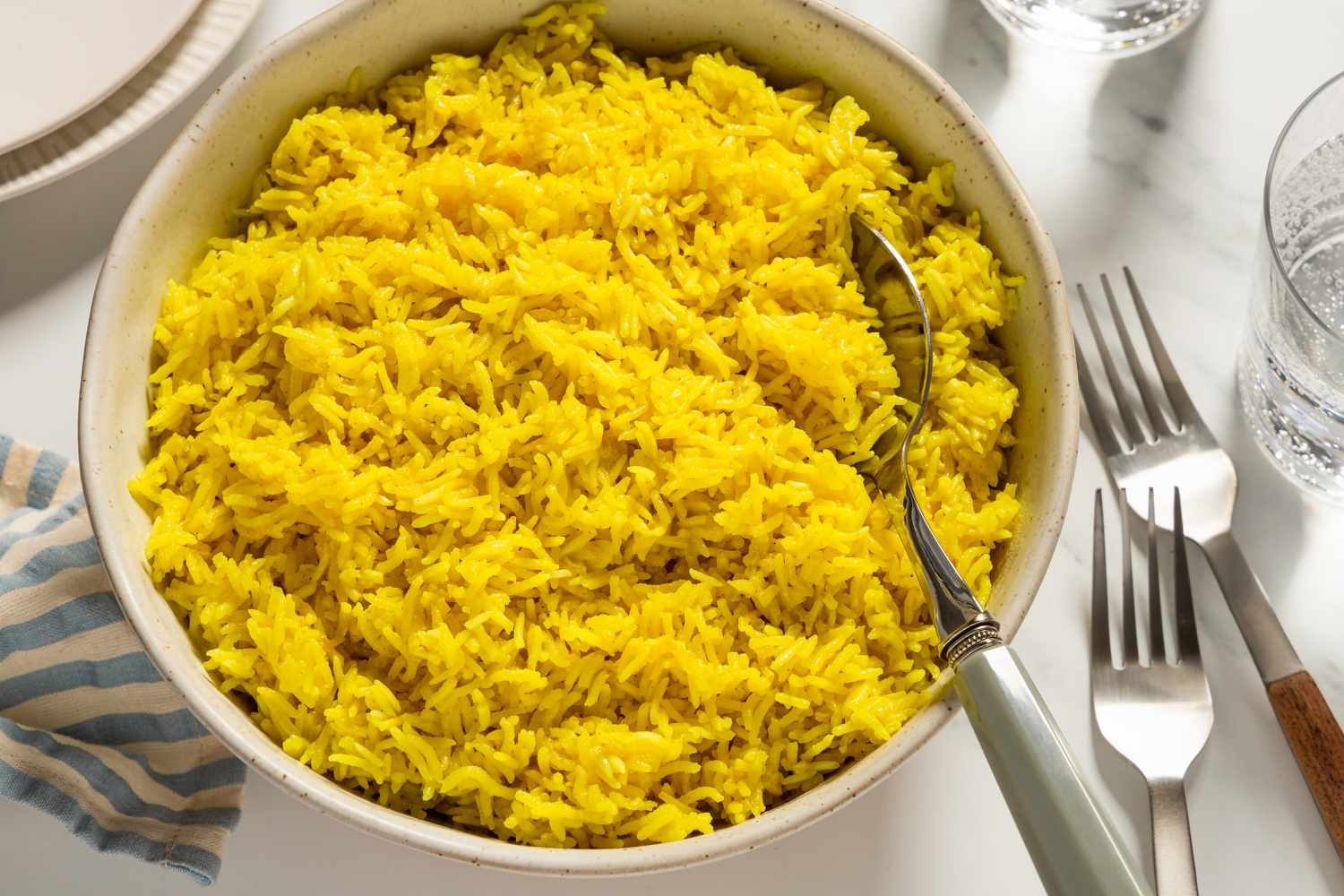Overhead view of a large bowl of golden rice with a serving spoon all on marble countertop with a cloth napkin, forks, plates a glasses