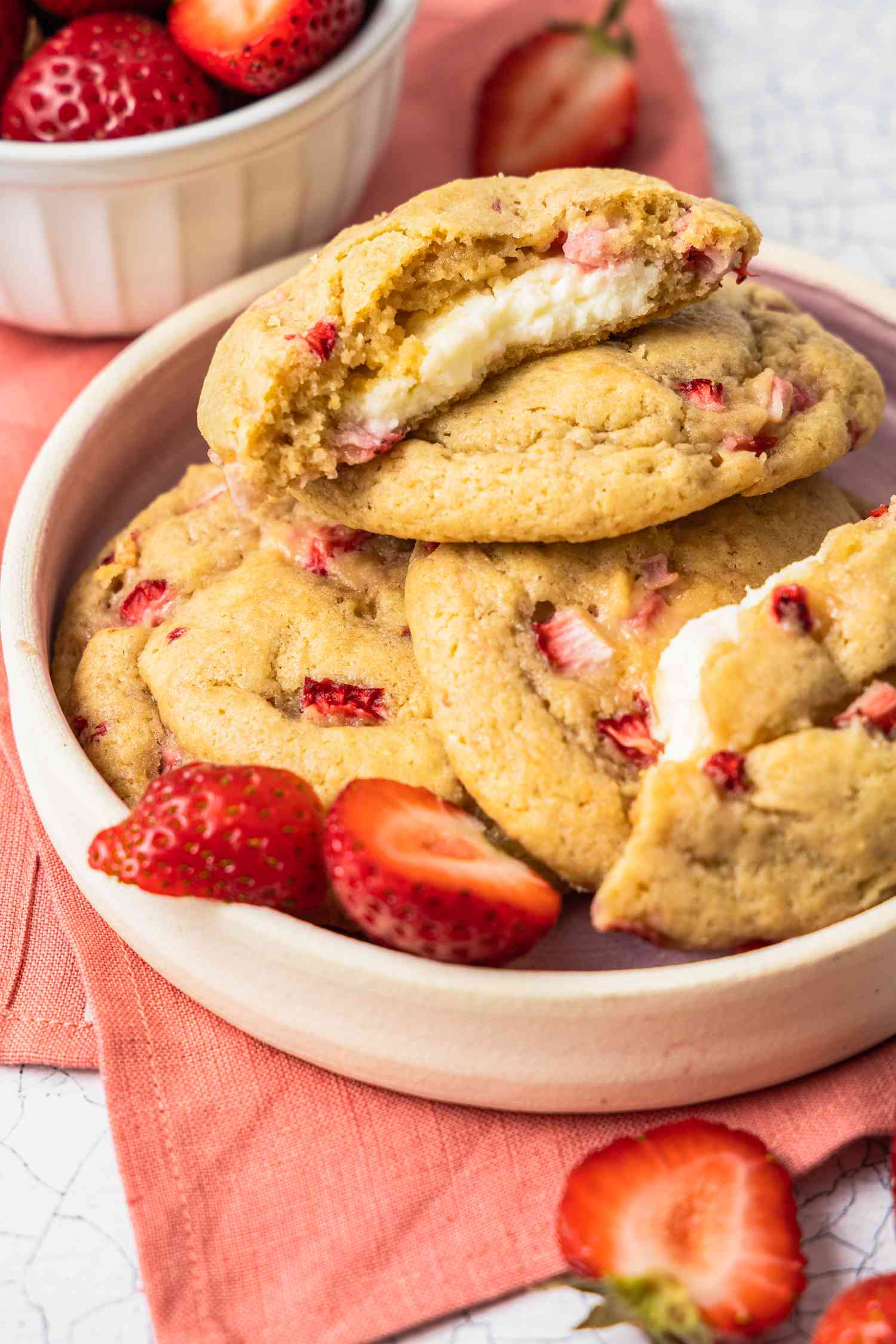Stack of Strawberry Cheesecake Cookies With the Top Cookie Cut in Half to Showcase Cheesecake Filling in a Small Plate With a Few Halved Strawberries, and in the Surroundings, a Bowl of Strawberries and Some Free Flowing Strawberries (Some Whole, Some Halved) on the Counter, All on a Pink Table Napkin 