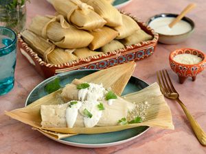 A Plate of Chicken Tamales with Chile Verde Topped with Crema, Cotija Cheese, and Some Cilantro at a Table Setting with a Platter of More Tamales, Utensils, a Small Bowl of Cotija Cheese, a Small Bowl of Crema, a Glass of Water, and a Small Vase