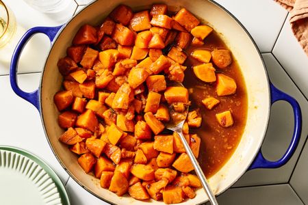 A pot of candied yams with a serving spoon viewed from above on a tiled surface with plate edges visible to the side