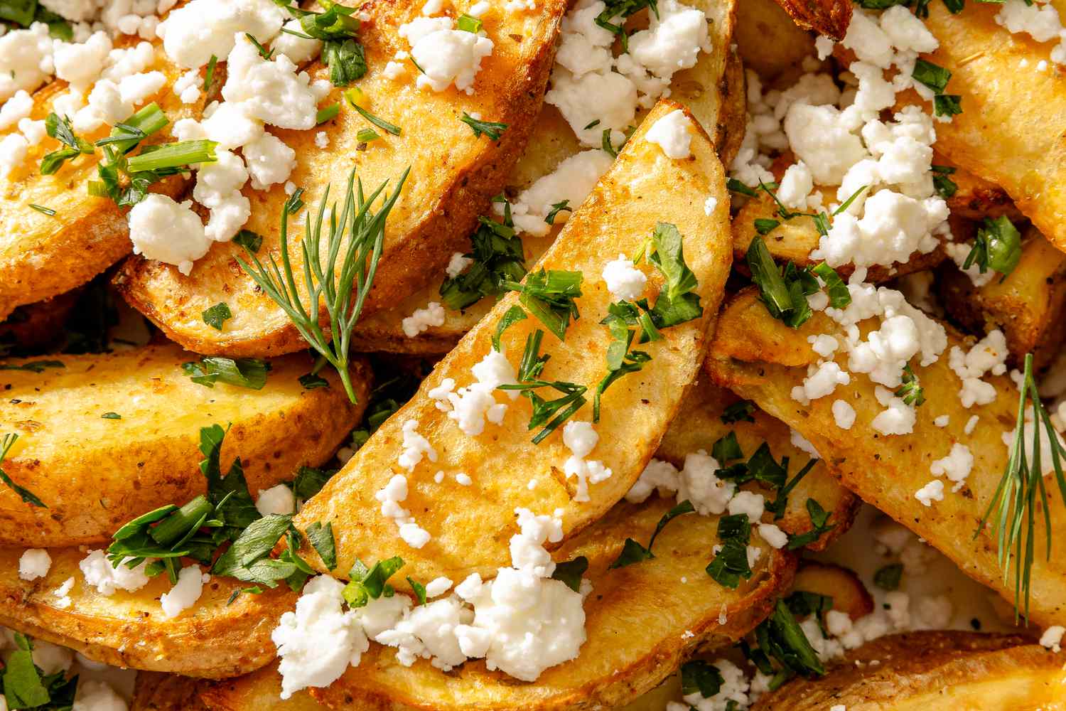 Closeup of Greek-style baked potato wedges, garnished with crumbled feta, dill and lemon wedges