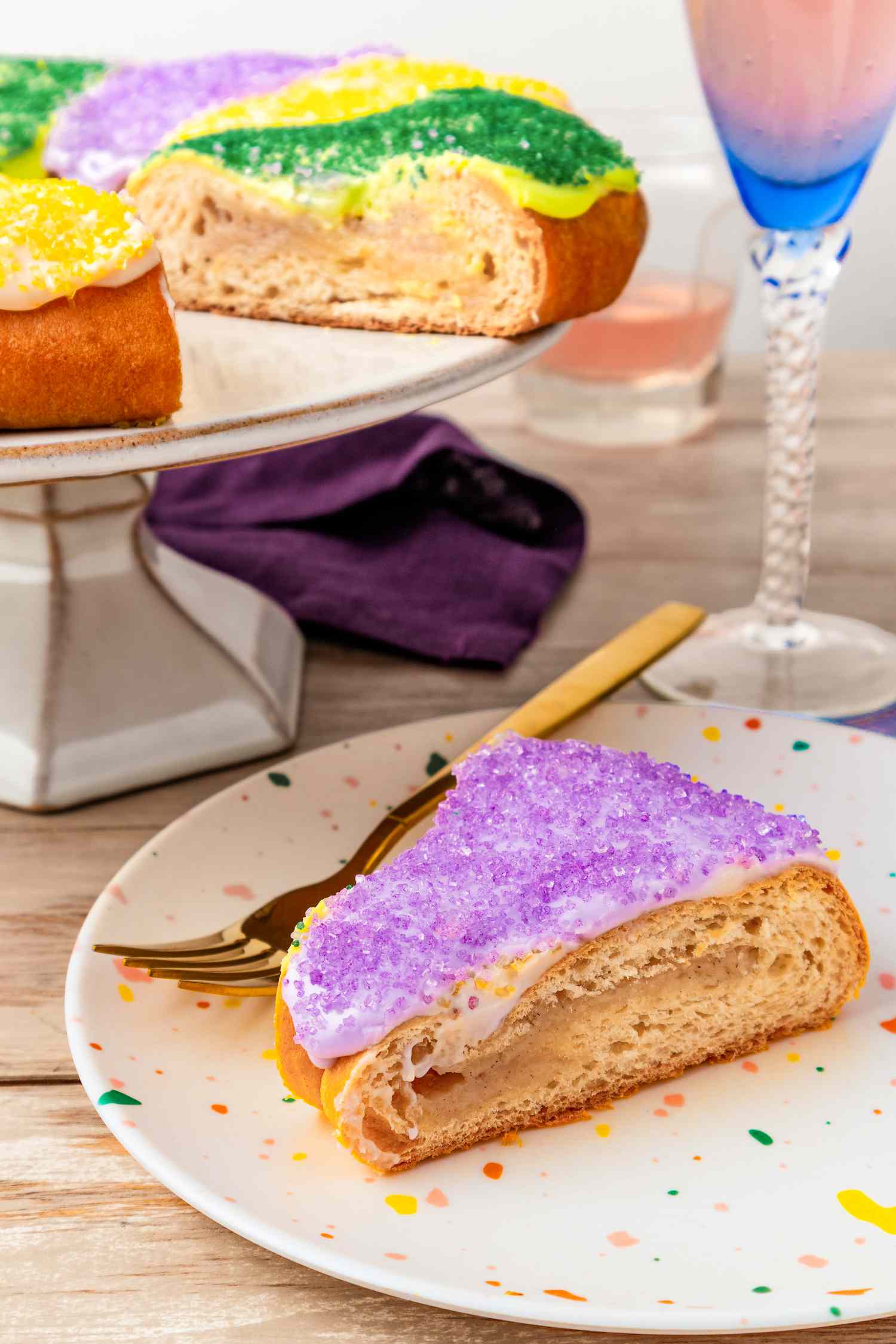 Slice of king cake on a plate with a utensil, and in the background, more on a cake stand at a table setting with drinks and a napkin