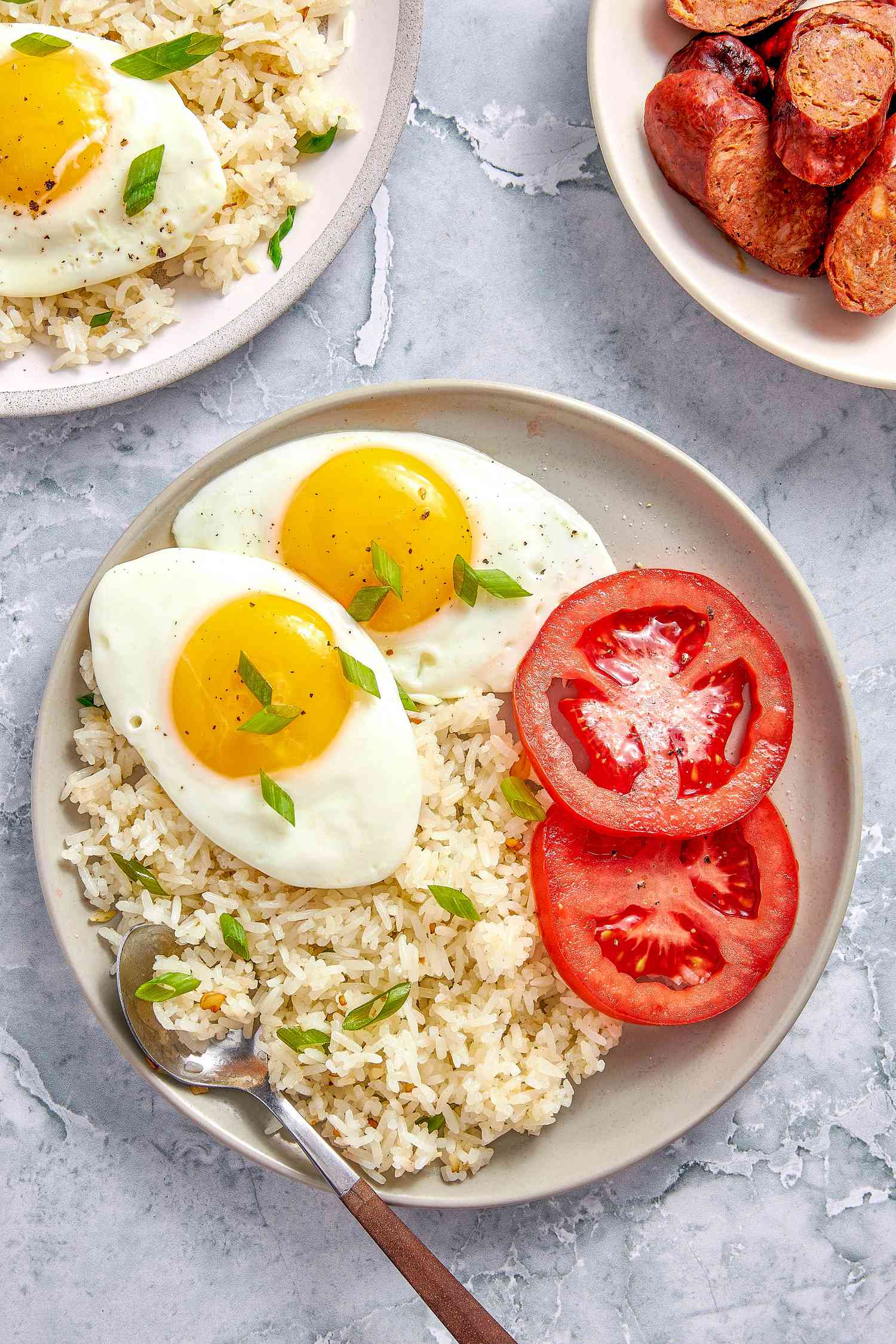 Plate of Garlic Fried Rice (Sinangag na Kanin) with sunny side eggs and sliced tomatoes, and in the surroundings, another plate with another serving of rice and eggs and a plate of longanisa (Filipino sausage)