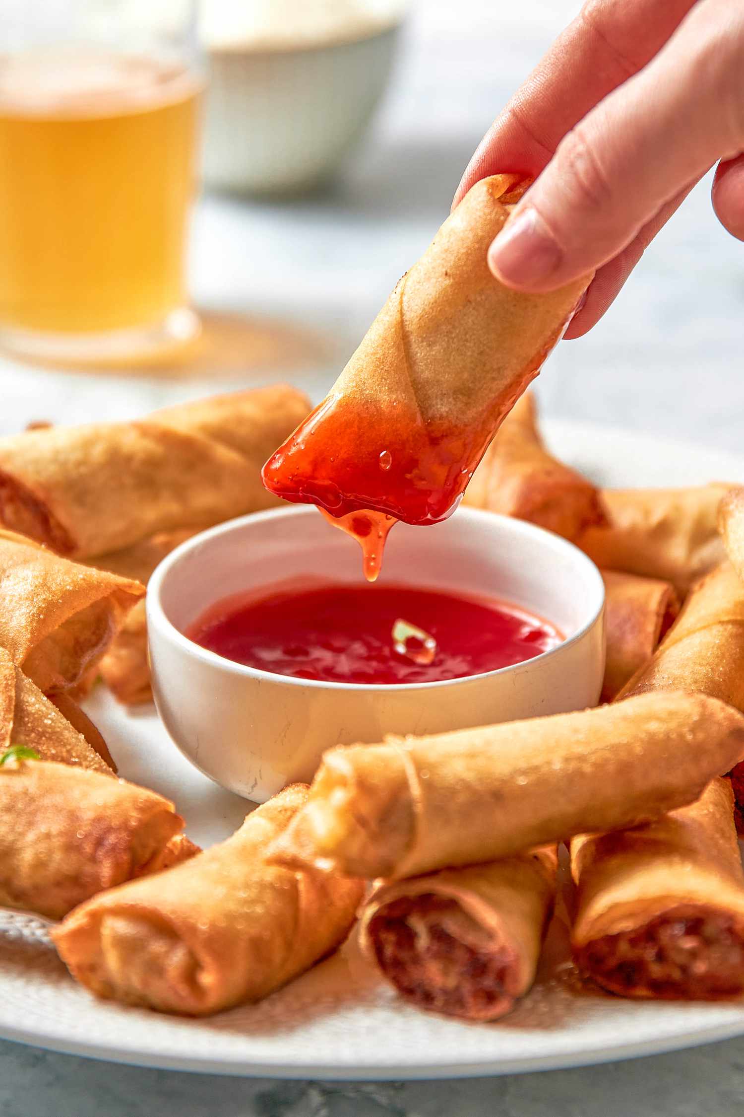 Piece of Lumpia Dipped in a Bowl With Sweet and Sour Sauce Sitting on a Plate With More Lumpia, and in the Background, a Glass With a Fizzy Drink