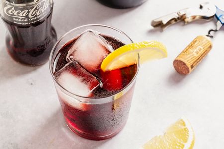 A glass of Kalimotxo with ice cubes and a lemon wedge, a cola bottle and a wine cork on the side