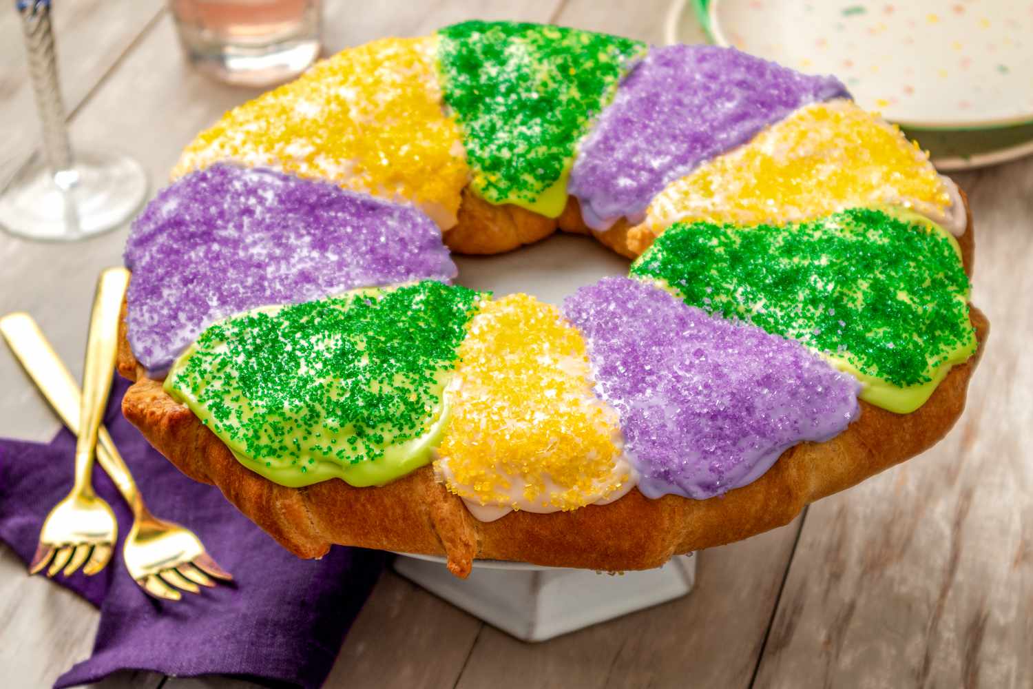 King cake (all decorated) on a cake stand at a table setting with utensils, a purple napkin, wine glasses, and a stack of plates