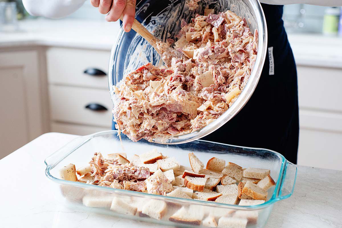 Reuben Casserole filling is being scooped onto the top of the chopped bread in the bottom of a glass casserole dish. The background is a kitchen and a marble topped island.