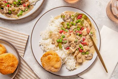 Serving of chicken a la king on a plate with rice and a biscuit at a table setting with another serving on a plate, a bowl of biscuits, glasses of wine, and table napkins