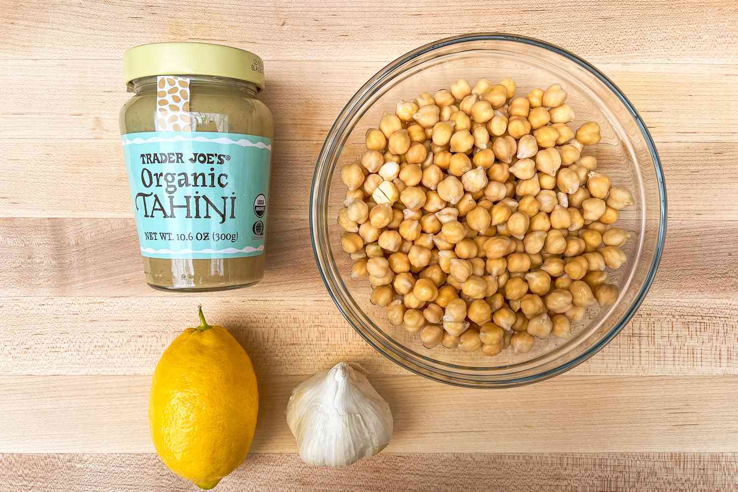 Jar of tahini, bowl of chickpeas, a lemon, and a garlic clove arranged on a wooden surface.