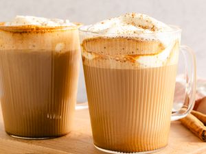Two mugs filled with latte topped with whipped cream sprinkled with spices and placed on a wooden surface