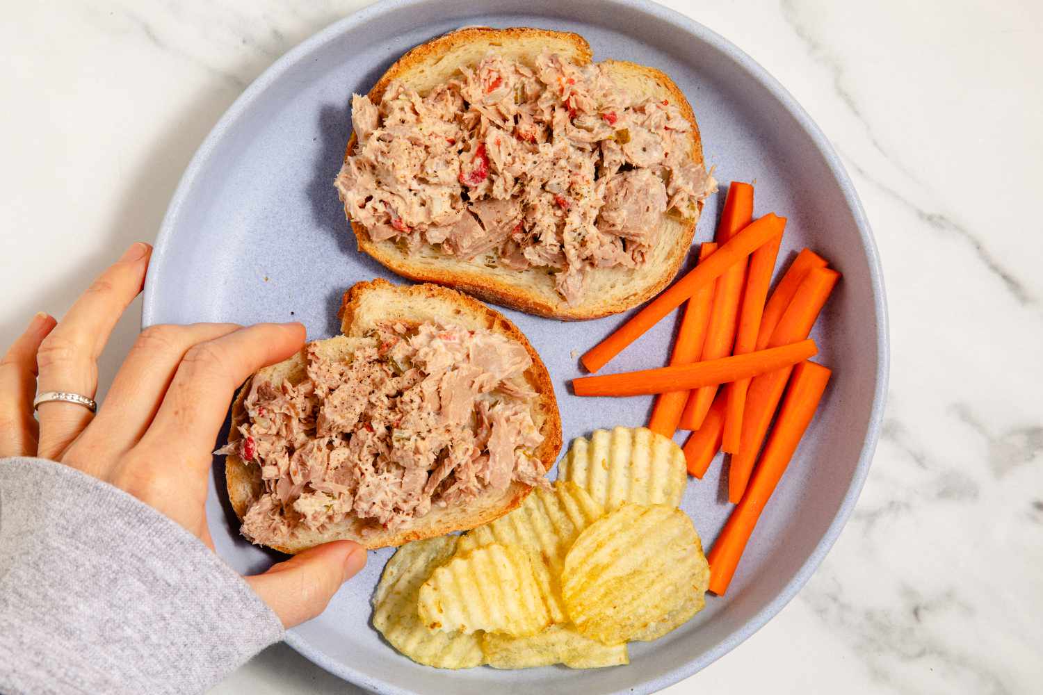 A hand grabbing one of two slices of bread with tuna salad, next to carrot sticks and potato chips on a plate