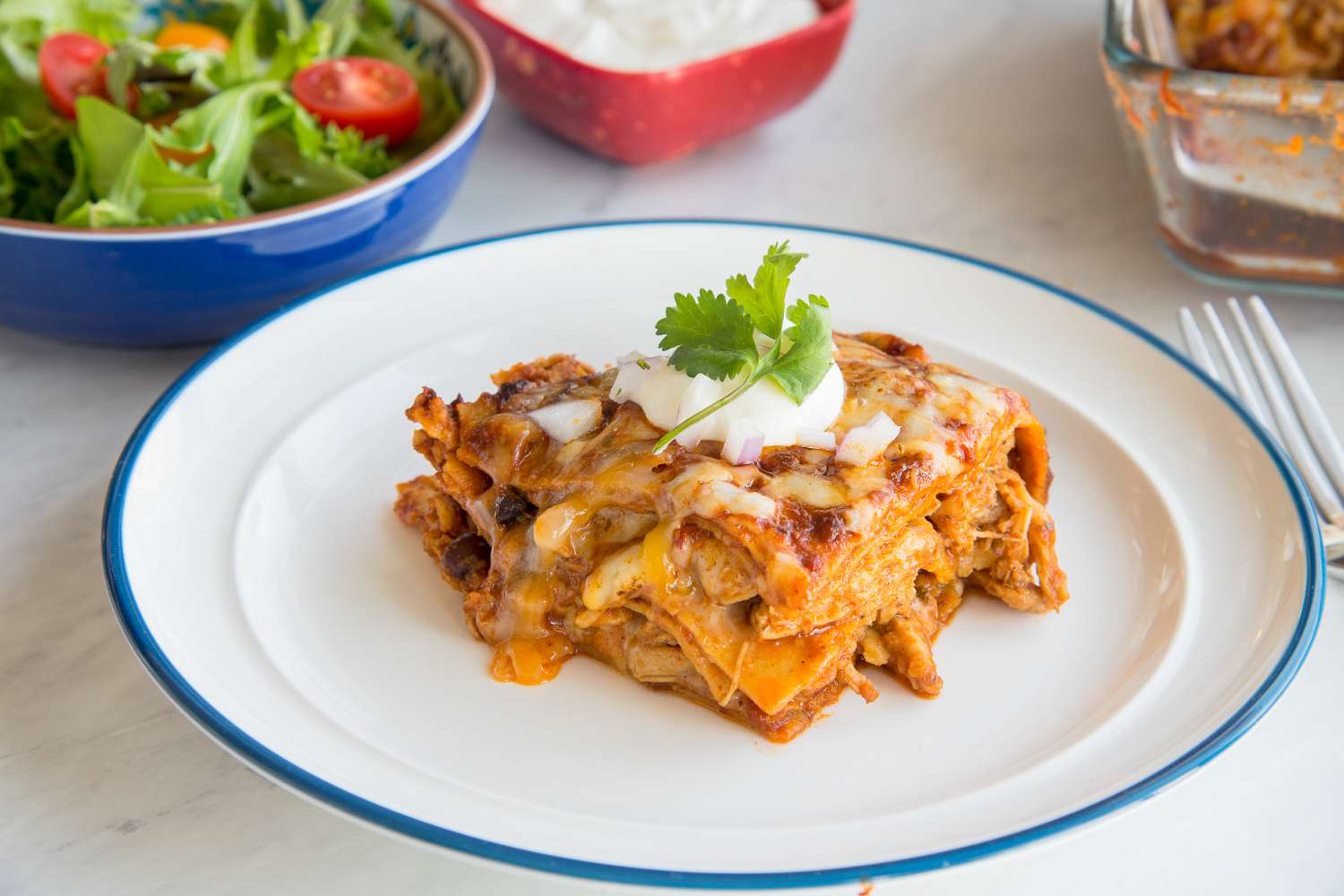 Slice of Easy Chicken Enchilada Casserole on a Plate Topped with a Dollop of Sour Cream, Some Onions, and Cilantro, and in the Background, a Bowl of Side Salad and Smaller Bowls with More Toppings