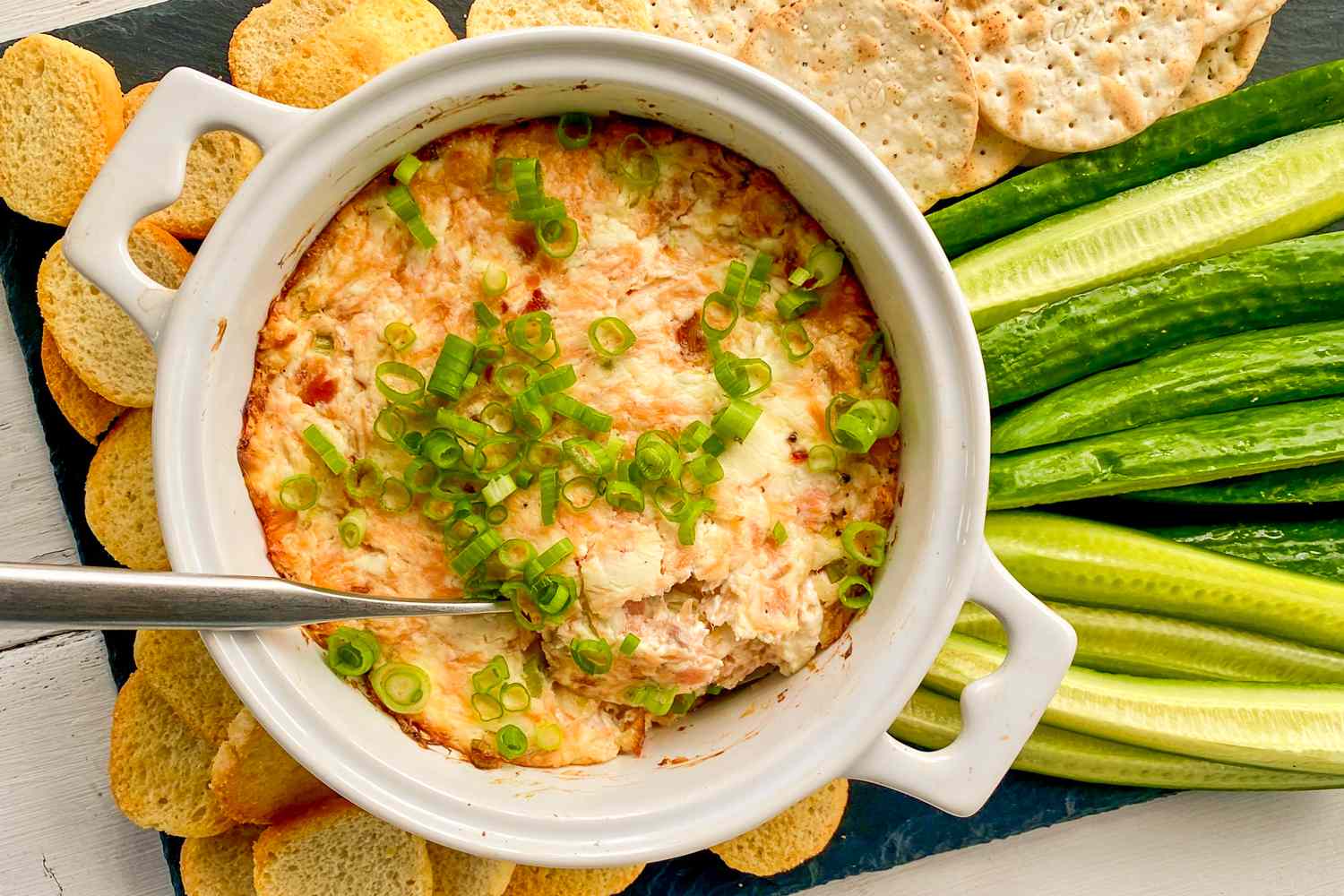 Overhead view of 3-ingredient smoked salmon dip in a white bowl topped green onions and surrounded by crackers, baguette slices and sliced cucumbers