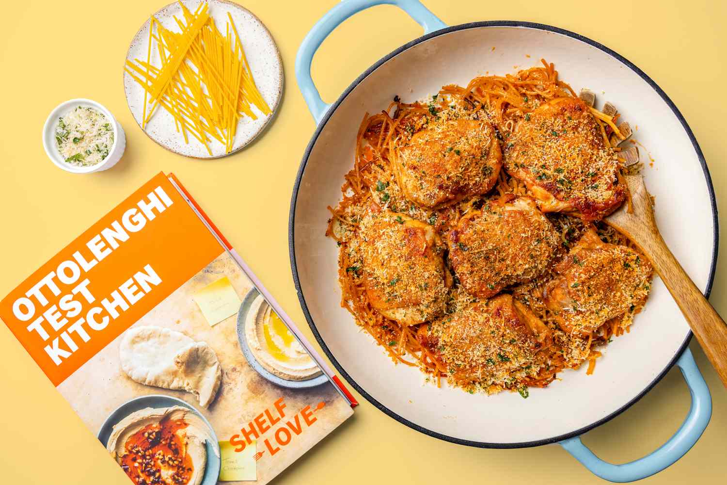 Dutch Oven With Ottolenghi One-Pot Spaghetti, a Plate With Dry Spaghetti Pieces, a Small Bowl of Breadcrumb Topping, and the Ottolenghi Test Kitchen: Shelf Love Cookbook, All on a Deep Yellow Background