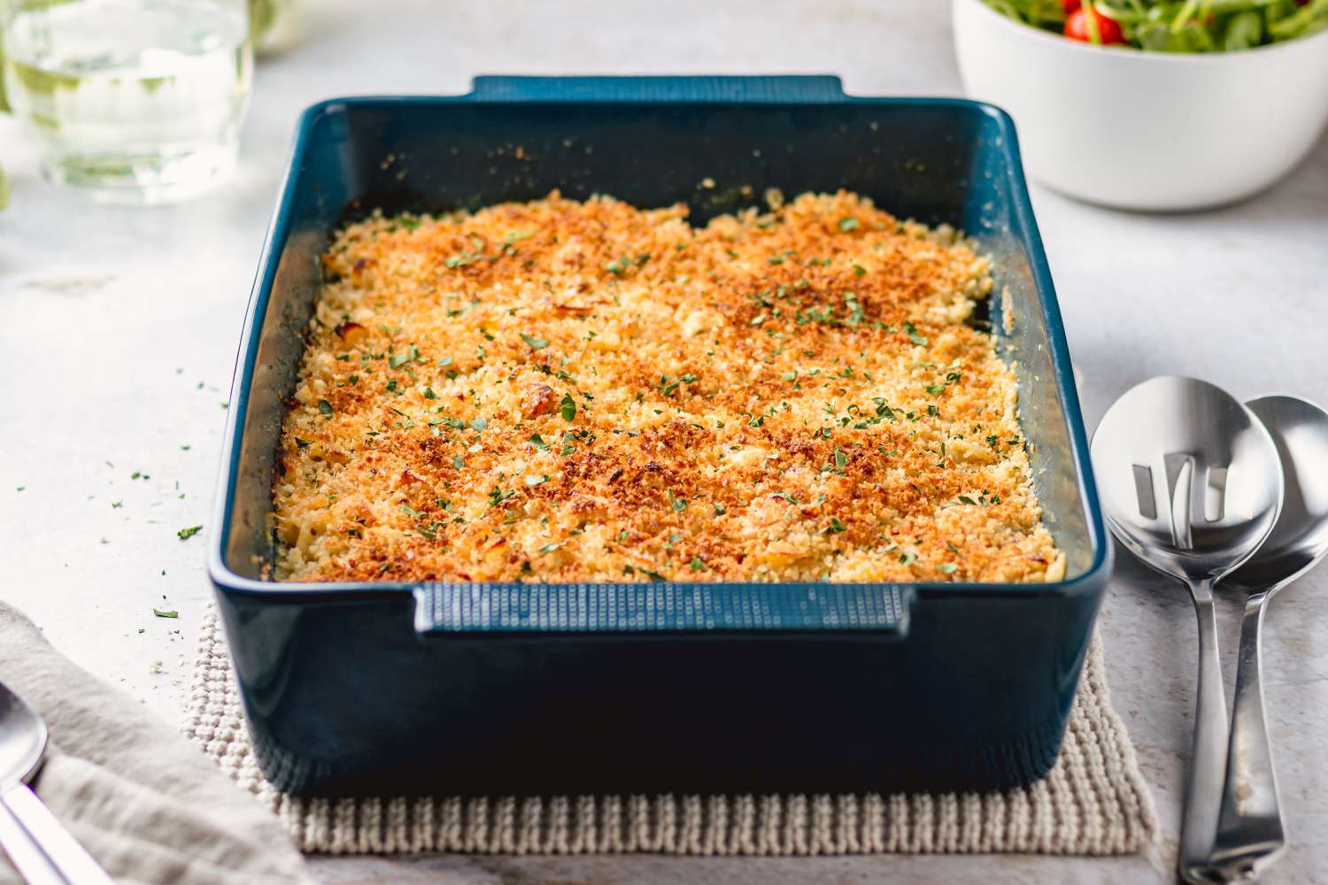 Angled view of a dark blue casserole dish with a casserole topped with crushed crackers next to two serving spoons, a glass of water and a bowl of salad greens