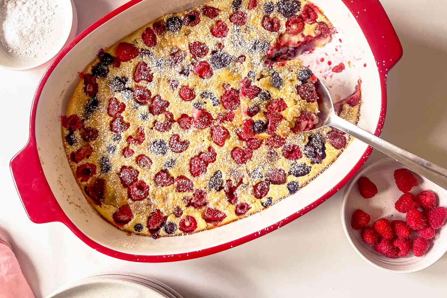 A berry-filled dish of clafoutis in a baking pan surrounded by bowls of fresh berries