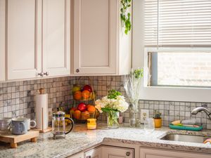 Photo of a clean kitchen, showing a kitchen sink with flowers, a basket of fruit, and a French press on the counter beside it