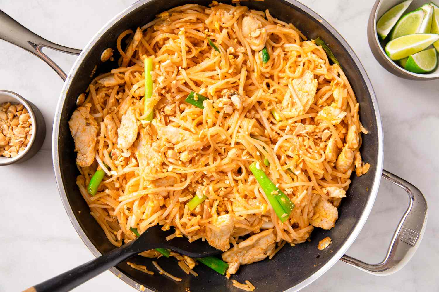 A pan of prepared pad thai with noodles vegetables and a serving utensil accompanied by small bowls of peanuts and lime wedges