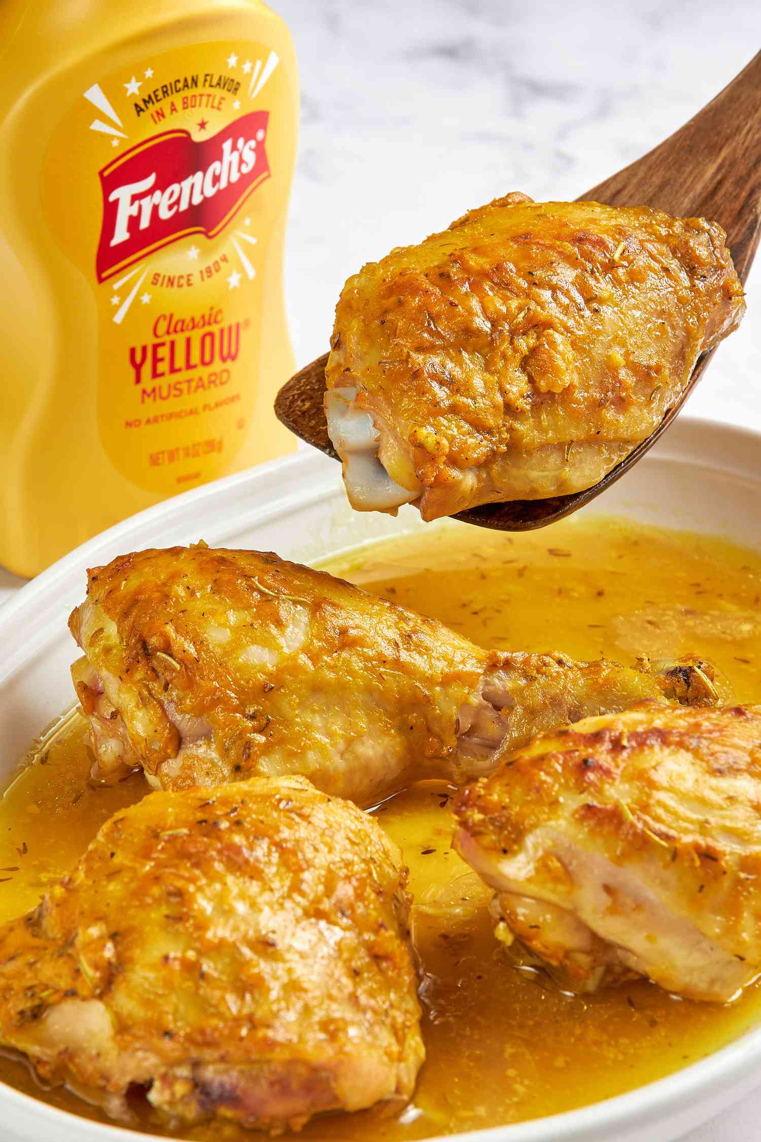 French's Mustard Chicken on a platter with one piece lifted on a spoon, and next to the platter, a bottle of French's Yellow Classic mustard