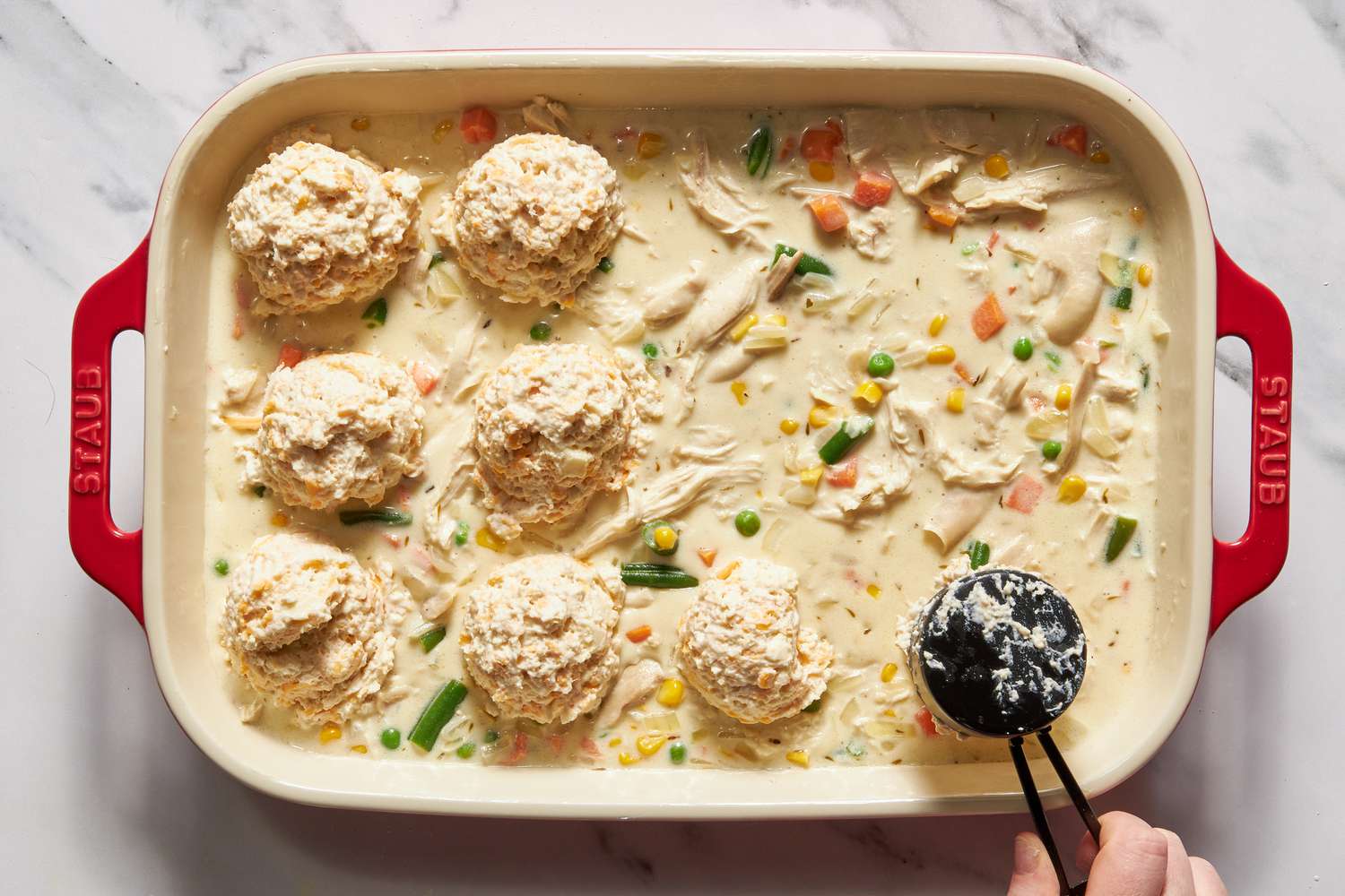 Dough spooned onto the casserole filling in the dish using a 1/4 measuring cup for chicken cobbler with cheddar bay biscuit top recipe