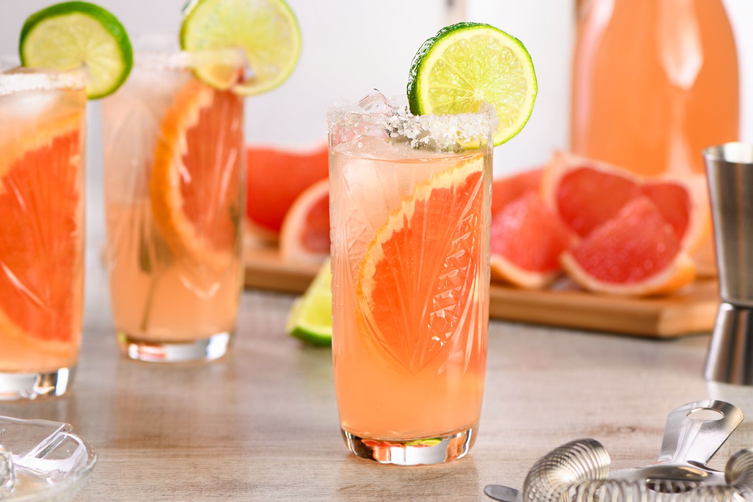 Paloma cocktail in a highbakk glass with a lime wheel garnish and cocktail ingredients in the background