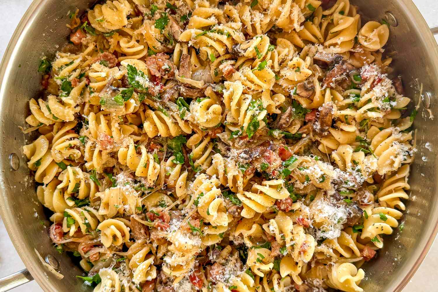 A skillet containing pasta with bacon mushrooms and grated cheese garnished with herbs